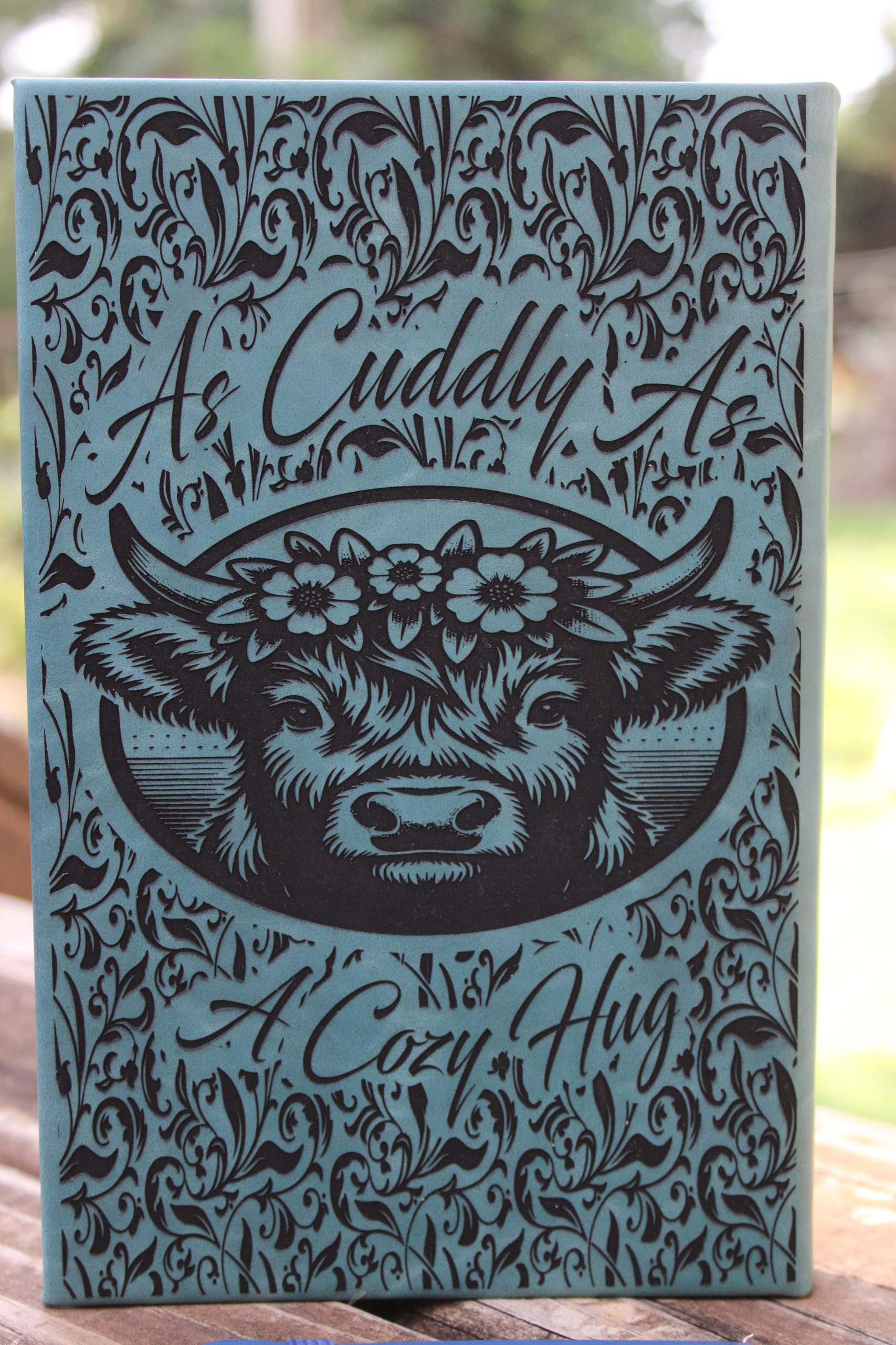 a teal journal with a highland cow and text reading 'as cuddly as a cozy hug' around the image. and a western tooled design surrounding the whole image