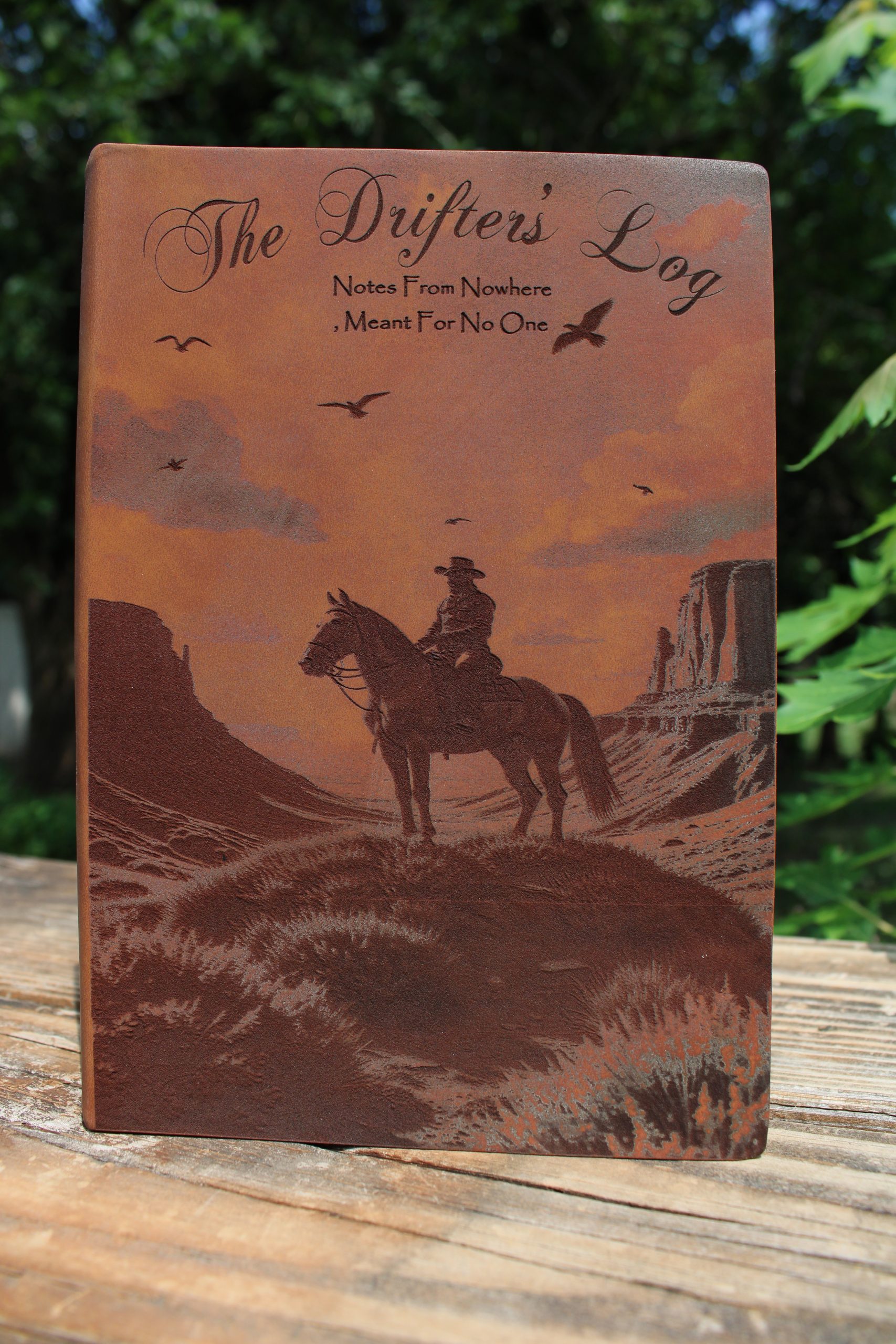 Brown soft touch journal with a cowboy riding a horse on top of a hill captioned 'the drifter's log'